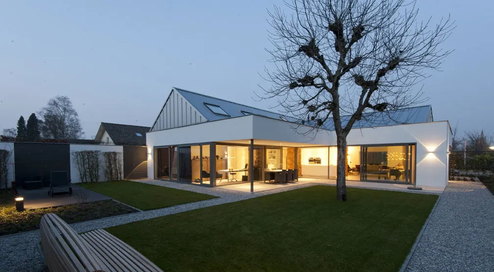 Dezelfde woning na de verbouwing tot villa, met witte gevel, grote glaspuien en zinken dak, bij avondlicht